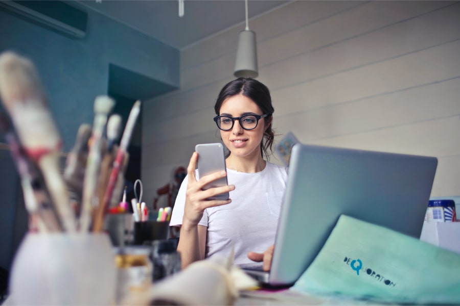 Uma jovem em frente a um computador portátil sorri enquanto consulta o seu telemóvel - A importância de uma marca comunicar bem