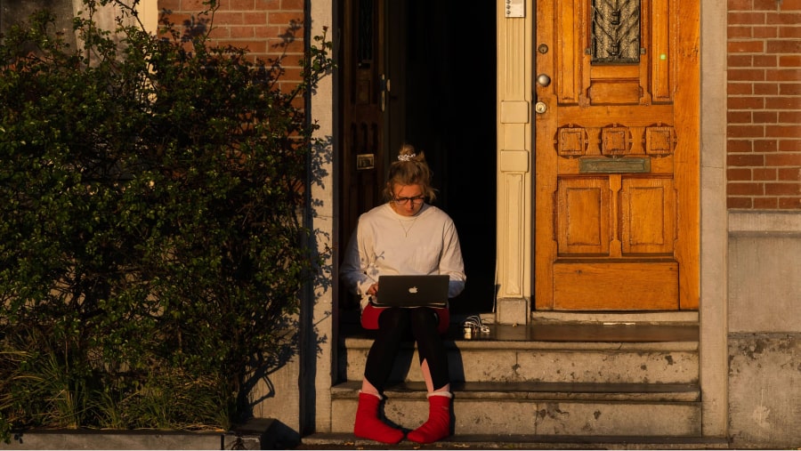 Mulher a trabalhar ao computador portátil sentada à entrada de casa, num dia de sol - Marketing Digital Home Office.